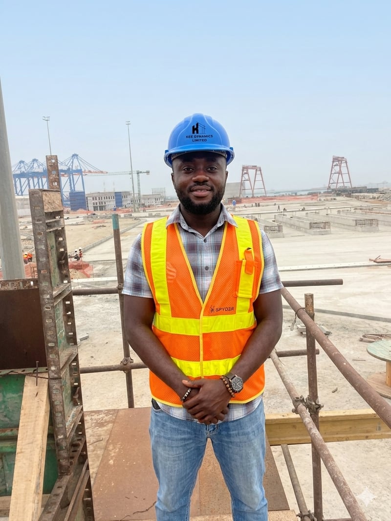 Kwame Owusu-Ansah wearing a blue KCE Dynamics Limited hard hat and high-visibility safety vest, standing at a large-scale construction site with cranes and concrete foundations under a clear sky.