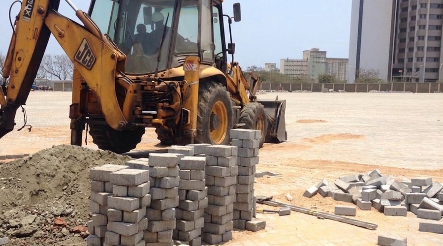Backhoe loader and stacked concrete paving blocks on the construction site of the GPHA Transit Terminal, Tema, with the city skyline in the background.