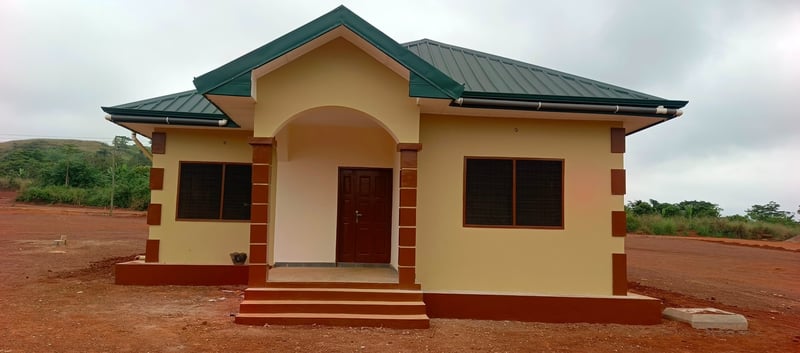 Completed single-story residential bungalow with cream walls, maroon trim, and a green metal roof, built on a red-earth lot in Ghana.