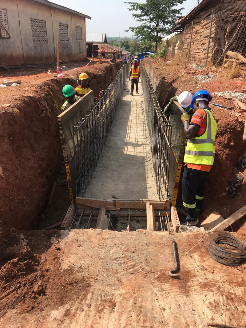Construction workers in hard hats and safety vests building a reinforced concrete retaining wall in a deep excavation trench, with rebar cages and timber formwork visible.