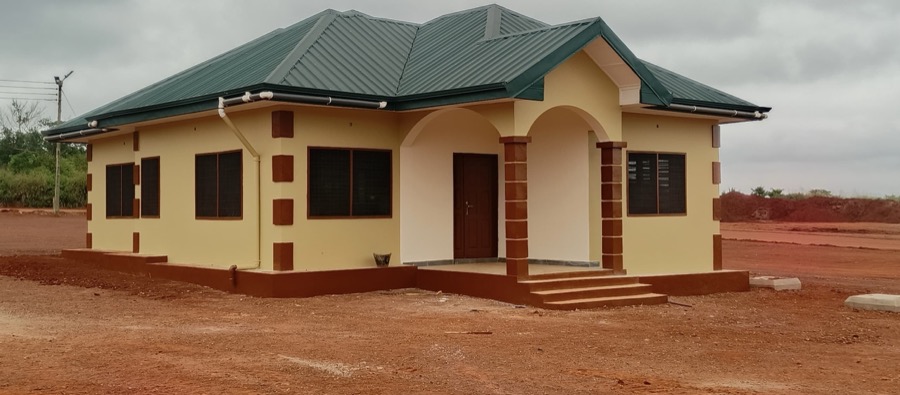 Completed resettlement bungalow with cream walls, maroon trim, and a green metal roof, Ahafo Region, Ghana.