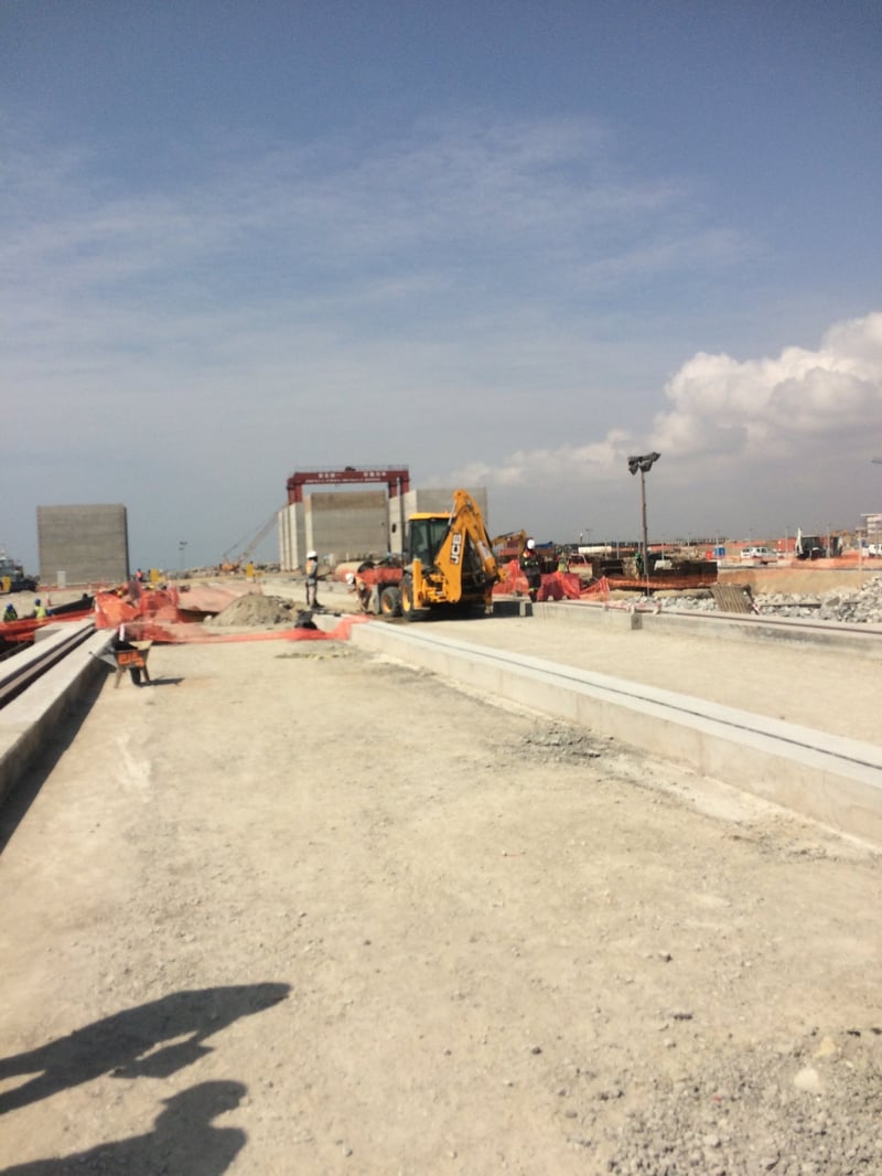 Wide view of a large-scale construction site with a backhoe loader, a concrete gantry structure, and workers at a coastal port development under a clear blue sky.